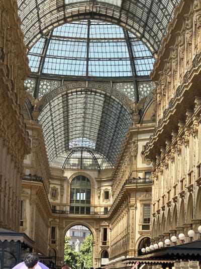 The image captures the Galleria Vittorio Emanuele II in Milan, Italy. This historic shopping arcade features an ornate glass and iron roof, supported by intricate metalwork and arches. The walls of the arcade are lined with elaborate stonework, including columns, statues, and balconies. Sunlight streams through the glass ceiling, illuminating the interior. In the foreground, the tops of awnings and some people are visible, suggesting a public space with commercial activity. Through an archway further down the arcade, a statue and trees can be seen, indicating an outdoor area beyond. The overall impression is one of grandeur and architectural splendor.