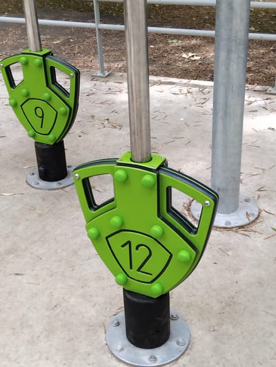 The image shows two green exercise equipment components, each attached to a vertical metal pole. The component in the foreground is a shield-shaped piece with the number "12" prominently displayed in black. It has several green spherical knobs on its surface and black trim around its edges. This component is mounted on a black cylindrical base. In the background, a similar shield-shaped component with the number "9" is visible, also attached to a metal pole and mounted on a black base. Both bases are secured to the ground with metal plates and bolts. The ground surface is concrete, with scattered dry leaves and debris. Behind the equipment, a metal railing is visible, and beyond that, a grassy area with trees. The scene appears to be an outdoor exercise park. The image does not provide enough information to determine the specific location in Vila Nova de Famalicão, Portugal.
