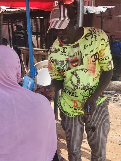 The scene shows a man in Kwamba, Nigeria, interacting with a person wearing a pink hijab. The man wears a yellow Chanel-patterned shirt, horizontally striped pants with a hole, and a patterned cap. He holds a mobile phone in one hand. In the background, there is a partially constructed wooden structure and some visible urban elements. The ground appears to be sandy or dusty.