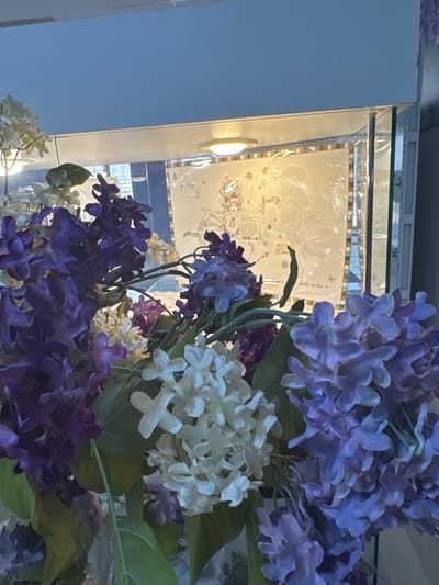 The image depicts a display inside a glass cabinet or shelf, likely in a retail setting or museum. The background features a decorative pattern with drawings of what appear to be Alice in Wonderland characters. The main subject consists of artificial flowers, primarily in shades of purple and white. The location is potentially in Klin, Russia, as the Alice in Wonderland theme is known to be present there, but it's not verifiable from the image alone.