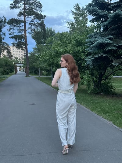 A woman with long, wavy reddish-brown hair and glasses stands on an asphalt path in an outdoor park or green urban area in Kazan, Russia. She is wearing a light-colored, vertically striped sleeveless jumpsuit with a belt, and light-colored high-heeled sandals. Her back is partially turned to the camera as she looks down and to her left.

The setting features an asphalt path bordered by grassy areas and numerous trees. On the left, a tall pine tree with a prominent trunk stands, and dense blue-green conifers are visible in the upper part of the frame. Various green deciduous trees fill the background. Distant multi-story urban buildings can be seen through the foliage. The sky is partly cloudy, suggesting it is daytime, possibly late afternoon or early evening. No visible text is present in the image.