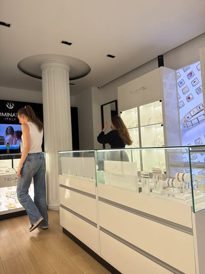 The image captures the interior of a brightly lit Nomination Italy jewelry store, likely located in Sofia, Bulgaria. The perspective is from an elevated angle, looking across the retail space.

Two women are visible. On the right side of the frame, a woman with long brown hair, dressed in a black top, is leaning into a glass display case, closely examining its contents. Her reflection is cast in a mirror situated behind the display. Towards the left, another woman with brown hair, tied back, wearing a white t-shirt and blue jeans, stands facing a different display case, looking down at the showcased items.

The store is furnished with modern, white display cabinets featuring glass tops and fronts. These cases are filled with various jewelry pieces, including bracelets, rings, and charms, illuminated by internal lighting. The "NOMINATION ITALY" logo is clearly visible on multiple display cases and possibly on a digital screen in the background. The flooring consists of light-toned wooden planks.

A prominent architectural feature is a large, fluted white classical column with a circular base and capital, extending from floor to ceiling in the central left area of the image. The ceiling is white and incorporates recessed lighting fixtures. A small black security camera is mounted high on a wall. The repeated brand name "NOMINATION ITALY" is evident, and faint Cyrillic text can be observed on some product labels or informational signs within the display cases, supporting the location context of Sofia, Bulgaria. The scene depicts a typical retail environment during what appears to be business hours.