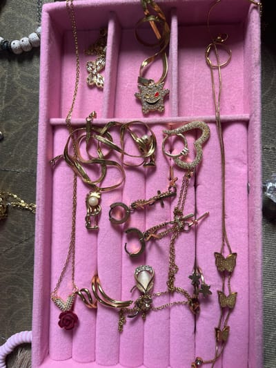 The image displays a pink, felt-lined jewelry organizer containing various gold-colored pieces, situated in the city of Ipetumodu, Nigeria. The organizer features multiple compartments.
**Objects within the organizer:**
- **Necklaces:** Multiple gold-colored necklaces are present. One necklace features a butterfly pendant encrusted with clear stones. Another has a Gengar-shaped pendant, detailed with clear stones and red eyes. A different necklace displays two brushed gold-colored butterfly pendants. Further necklaces include a pendant of a stylized figure holding a white pearl, a large heart-shaped pendant covered in clear stones, a red rose pendant beneath a V-shaped element with clear stones, an oval "cat's eye" stone pendant framed by clear stones, and a long chain with multiple small brushed gold-colored butterfly pendants and small spherical beads. Two necklaces feature small cross-shaped pendants.
- **Earrings:** Two gold-colored hoop earrings are visible in one compartment.
**Surrounding Environment:**
- To the immediate left of the organizer, a segment of a beaded bracelet with white, textured spherical beads and dark spacers rests on a green fabric surface.
- To the right of the organizer, a brown fabric surface is visible, upon which a small, clear, irregularly shaped object is positioned.