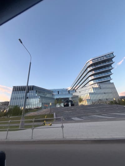 The image depicts the modern architecture of a business center in Moscow, Russia. The building features a distinctive wave-like design with multiple tiers of glass facades, reflecting the clear blue sky and soft sunset clouds. In the foreground, a wide staircase leads up to the building, with a black spherical sculpture positioned at its base. A yellow electric scooter is visible on a paved pathway near the stairs. A street lamp with a curved pole stands to the left of the frame, and a metal fence runs along the foreground, separating the pathway from a road. The overall scene suggests a contemporary urban environment, likely during the late afternoon or early evening.