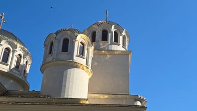 The image shows the domes of the Dormition of the Mother of God Cathedral in Varna, Bulgaria, against a clear blue sky. The cathedral's domes are gilded and white, with arched windows. Sunlight reflects off the golden elements, creating bright highlights. One of the domes has a cross on top. In the upper left corner of the image, a small bird is visible in flight. The architectural style suggests a religious building, characteristic of Eastern Orthodox churches. The perspective is from below, looking up at the structures.