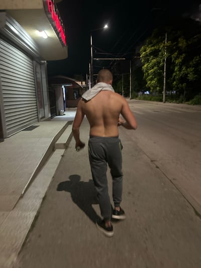 A shirtless young man with short hair walks away from the camera at night on a sidewalk next to a street. He is wearing dark gray sweatpants and black sneakers. A light-colored towel is draped over his shoulders. He is holding a small, clear object in his right hand. The sidewalk is made of concrete and appears to be slightly sloped. To the left of the sidewalk is a building with a closed metal shutter and a lighted sign with red lettering. Beyond the building, a small structure with an open window is visible. The street to the right of the sidewalk is dark and appears to be paved. Trees line the far side of the street. Overhead power lines are visible against the dark night sky. The scene is lit by artificial lights from the building and a street lamp. The overall impression is that of a quiet urban street at night.