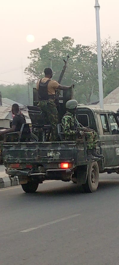 A military vehicle carrying armed soldiers drives on an asphalt road. The vehicle is dark green and has a large machine gun mounted on the back. Three soldiers are visible; one stands behind the gun, wearing a flak jacket and camouflage pants, another sits in the back, and the third sits in the cab, wearing a helmet. The sky is overcast, with the sun partially visible. Buildings and trees line the background.