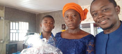 Three people are captured in a close-up shot inside what appears to be a building. On the left is a young boy holding what looks like a gift wrapped in clear plastic. In the middle is a woman wearing an orange head wrap and a blue lace dress. To the right is a man in a blue shirt, smiling. In the background, there's a patterned wall, a window with metal bars, and a speaker. A microphone on a yellow stand is partially visible behind the woman. The setting appears to be indoors, and the presence of a microphone suggests it might be a community or event space. The location is Lokoja, Nigeria, based on the cultural dress and potential community setting.