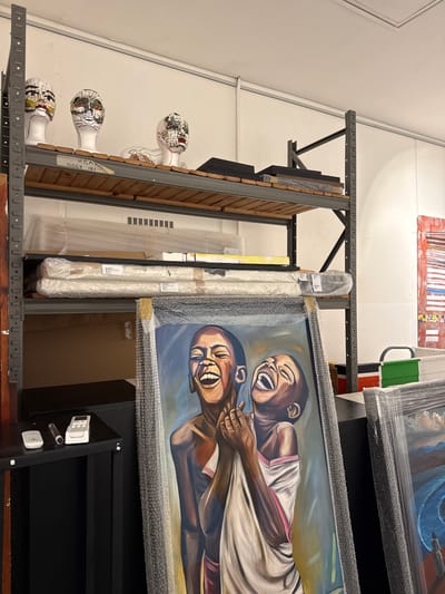 The image shows an interior storage space, possibly in an art storage facility, with artwork arranged on metal shelving. The artwork includes framed paintings wrapped in bubble wrap, wooden panels, and decorative mannequin heads. One painting prominently features two figures laughing. Other paintings are visible in the background. The exact location within Cape Town, South Africa, cannot be determined from the image.
