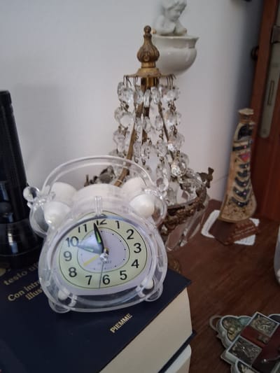 A close-up, eye-level shot captures a still life arrangement on a wooden surface, likely a table or shelf, within an interior. The foreground is dominated by a transparent, round alarm clock with white accents, its face displaying the time as approximately 10:56. The clock rests on a dark blue book with gold lettering, partially obscured by a stack of books underneath. To the left of the clock, a black flashlight stands upright. Behind the clock, a decorative, antique-looking lamp with numerous crystal pendants is the central focus. Further back, a small porcelain figurine of a child is perched on a white pedestal. To the right of the lamp, a tall, slender decorative item with a patterned design and a dark top stands. A sliver of a wooden door frame is visible on the far right. The background is a plain white wall. The overall scene suggests a domestic setting, possibly a study or living area. The image composition and the objects present do not provide any specific clues to its location in the city of Colorno, Italy.