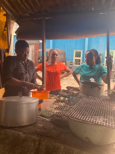 The image captures an outdoor or semi-outdoor food preparation scene, likely a market stall or street vendor in Aba, Nigeria. Three women are visible, engaged in various tasks under a rustic shelter with a corrugated metal roof section visible on the left.
In the foreground, a woman wearing a black long-sleeved top and a dark head covering is seated, attentively peeling or preparing a small root vegetable, possibly ginger, with a knife. A large silver pot with a lid sits beside her. Behind her, another woman in a vibrant orange top stands with hands on her hips, looking upwards towards the camera with a neutral expression.
Further back and to the right, a third woman wearing a teal t-shirt stands, looking down at a large pot or container, possibly stirring its contents or attending to it. She has braids. To her right, a large metal grate or rack is prominent, with various items, including another pot, visible around it, suggesting a cooking or grilling area. An orange bucket is also visible on the ground.
In the background, a bright blue building with white-framed windows stands under what appears to be a clear, sunny daytime sky. Part of a vehicle, possibly a tricycle (keke), is visible between the two standing women, with text "A5500" and "ASOPU" partially legible on its side. The ground appears unpaved, adding to the informal, bustling atmosphere of a street food setup. The overall mood suggests focused activity in a lively environment.