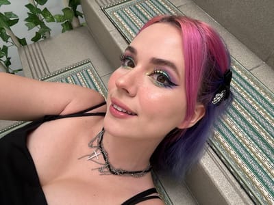 A sideways, eye-level close-up shot features a woman with pink and purple hair and a complex eye makeup look, lying on a textured surface that appears to be a rug or mat. The woman wears a layered black choker necklace with cross charms and a black top. Some artificial green leaves are partially visible on the right side of the frame. The image contains no information about the City of Zagreb, Croatia.