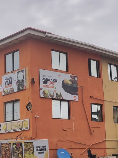 The image shows a close-up of the exterior of a two-story building painted orange and yellow. Several windows with dark frames are visible. On the orange section of the building, there are two large billboards. The left billboard advertises "POUNDED YAM" and displays images of food. The right billboard says "AMALA ON THE SPOT" and also features images of food. Below these billboards, there is another smaller sign with images of various food items like fruit juice, yoghurt, shawarma, ice cream, and jollof rice. Further down, a portion of another large billboard is visible with text that reads "SATISFY YOUR CRAVINGS TODAY!". A blue satellite dish is partially visible at the bottom right of the image. The sky is overcast. There are no people visible in the image. This building appears to be a commercial establishment, likely a restaurant or eatery, located in the city of Jos, Nigeria, given the context of the food items advertised.