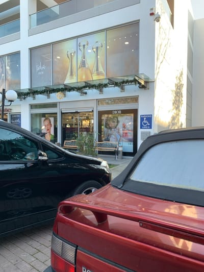 The image depicts a street scene in Marousi, Greece, featuring several high-end commercial storefronts and parked cars under bright, sunny conditions. The perspective is from slightly above, looking towards the building facade.
In the immediate foreground, a red car, possibly a sedan or coupe, is parked, showing its rear and right side, with some visible wear on its paint. Adjacent to it and further into the scene, a black car is parked, with its driver's side mirror and front wheel visible. Both cars are on a paved area, which appears to be a street or parking lot.
The background is dominated by a modern, multi-story building housing various luxury brand stores. The upper-level windows feature large graphics of elegant perfume bottles and the text "J'adore DIOR." Below this, a prominent "DIOR" store entrance is visible, accompanied by a large window display showcasing a woman's portrait, likely for perfume or makeup advertising. Below the Dior store, another entrance can be seen with the name "ESTEE LAUDER." Further to the left, a "LANCÔME" sign is visible above another display featuring a woman's face. The building's facade is decorated with green garlands, possibly holiday lights, and spherical decorative lights.
Notable details include a blue sign mounted on the building near the Dior entrance, which reads "PARKING AREA" and includes the international disabled access symbol. A security camera is mounted above the Dior store entrance. Two outdoor benches are placed in front of the stores. The bright sunlight casts distinct shadows on the building, indicating a clear day. No specific human activity beyond parked cars is explicitly depicted, though a faint reflection of a person is visible in a car window.
Visible text includes: "J'adore DIOR," "DIOR," "ESTEE LAUDER," "LANCÔME," and "PARKING AREA" with the disabled access symbol.