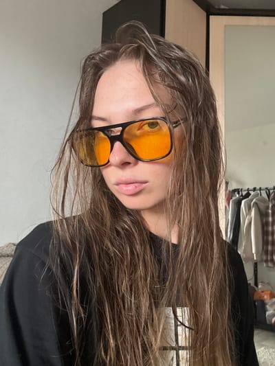 The image is a close-up, waist-up, medium shot of a young woman with long, brown, wet hair, wearing black sunglasses with orange lenses and a black t-shirt. She is looking directly at the camera with a neutral expression. The background appears to be an indoor setting, possibly a bedroom or dressing area, with a closet or wardrobe visible on the right side of the frame, containing hanging clothes. A portion of a wall is visible on the left, painted a light color. The overall lighting suggests an indoor environment, possibly natural light from a window or diffused artificial light. There is no identifiable indication of the location being in the city of Perm, Russia, or any specific urban setting.