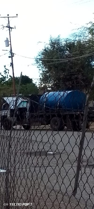 This is a medium-shot, slightly blurry, eye-level photograph taken outdoors during daylight, likely in Altagracia, Venezuela. The foreground is dominated by a chain-link fence, through which a scene unfolds.
Behind the fence, a dark-colored truck is visible. The truck is carrying a large, blue cylindrical tank. The cab of the truck appears to be white or silver, and the driver's side door is open. The truck is parked on a paved surface, which shows some cracks and debris.
In the background, a utility pole with a transformer and wires is present, suggesting an urban or semi-urban environment. Trees and foliage are visible further in the distance, with a bright, overcast sky above. The overall impression is that of a functional, possibly industrial or agricultural, setting. There are no people clearly visible in the frame.