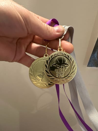 A hand is holding two gold medals with purple and gray ribbons. One medal features a basketball hoop and ball, while the other medal has a laurel wreath design. The medals are suspended from a light-colored wall. The image does not contain information about the city of Tallinn, Estonia.
