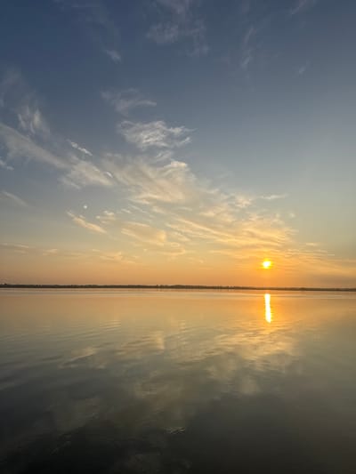 This is a landscape photograph of a serene body of water at sunset. The sun, a bright orange orb, is low on the horizon, casting a golden reflection across the water. Wispy white clouds streak across a pale blue sky, transitioning to hues of orange and yellow near the horizon. The water's surface is calm, with subtle ripples reflecting the sky and sunlight. A faint line of trees or structures is visible on the distant shore, creating a subtle silhouette against the vibrant sky. The image evokes a sense of tranquility and natural beauty. The photograph does not depict any people, actions, or specific objects that would indicate a location within the city of Lyman, Ukraine.