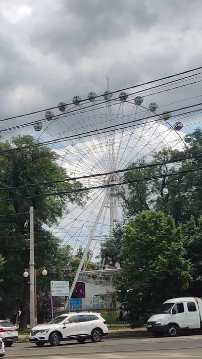 A white SUV and a white van are parked on a street in Krasnodar, Russia. In the background, a large Ferris wheel is visible behind trees and power lines. The sky is cloudy. A street sign is also visible, indicating street names in Cyrillic.
