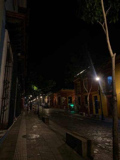 The image depicts a street scene at night in Oaxaca City, Mexico. The buildings are brightly lit with yellow and red paint. Along the sidewalk are lamp posts that illuminate the path. There are several people standing and walking along the street. The foreground of the image shows a series of metal railings.