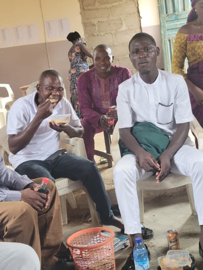 The image depicts an indoor gathering in Madalla, Nigeria. In the foreground, a man in a white shirt and pants is seated, looking directly at the camera. Beside him, another man in a white polo shirt is eating from a plastic container. To the left, partially visible, is another person in brown pants. In the background, a woman in a patterned dress is standing. Another man in a purple garment is seated. The environment appears to be a simple room with cinder block walls and a light-colored floor. Objects visible include plastic chairs, a bottle of dark liquid, a can of malt drink, books, a plastic basket, and food containers. The scene suggests a casual meal or social event.