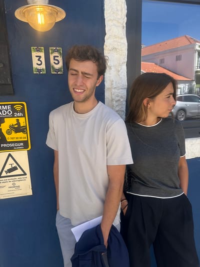 An outdoor portrait captures two individuals standing against a vibrant blue building facade under bright daylight, likely in Torres Vedras, Portugal, given the visible Portuguese text. The man in the foreground, wearing a light-colored t-shirt, smiles broadly with his eyes closed, showing a joyful expression. He has short, curly brown hair and light stubble. His left arm is visible, draped with a dark blue or purple item.
Behind him, a woman with long brown hair wears a grey t-shirt with white trim and black pants. Her eyes are also closed, and her expression appears serene and peaceful.
The blue facade features a metallic outdoor light fixture with an Edison-style bulb, and two decorative white tiles with blue floral designs, displaying the numbers "3" and "5". Below these, two security signs are mounted. The yellow sign shows a motorcycle icon and reads "ALARME ADO 24h SEGURANÇA", with a phone number "707 22 23 22" and "PROSEGUR ALARMES". The white sign states, "Para sua proteção, este local é objeto de videovigilância.", also featuring "PROSEGUR ALARMES" and "Alvará 248 A) C) do MAI", alongside a CCTV camera icon.
Above the blue wall, a textured white stone or stucco wall separates it from a large window reflecting a bright blue sky and the upper part of a building with a distinct red tiled roof. A partial reflection of a silver car is also visible in the window. The overall scene suggests a sunny day, with the individuals enjoying the light or posing for a photo.