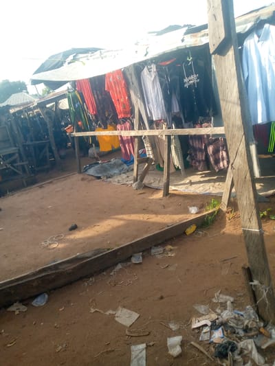 This image depicts an outdoor market scene in Jalingo, Nigeria. The setting appears to be a makeshift stall with a corrugated metal roof and wooden supports, sheltering an array of clothing items hanging on display. A variety of garments are visible, including shirts, t-shirts, and what appear to be trousers or skirts. Some of the clothing is brightly colored, such as an orange shirt with a red pattern and a yellow garment, while others are more muted, like white and light blue shirts, and dark colored t-shirts. One black t-shirt has white lettering, which reads "MADNESS NEIGHBORHOOD". The ground is covered in sandy soil, with scattered debris including plastic bags, wrappers, and some unidentifiable objects. In the background, there are more stalls and structures, suggesting a larger marketplace. The lighting indicates it is daytime, with sunlight casting shadows on the ground. The overall impression is one of a bustling, yet somewhat unkempt, open-air market. There are no people directly visible in the foreground or midground, but their presence is implied by the commercial activity.