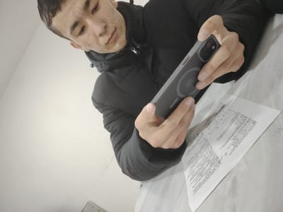 An overhead, slightly angled photograph captures a young male in Kungrad, Uzbekistan, observed from a low perspective. The individual, appearing to be in his late teens or early twenties, with short dark hair and visible facial blemishes, is wearing a black puffer jacket. He is intently focused downwards, holding a dark grey smartphone with a MagSafe-compatible case in both hands. The phone's camera array is visible. Positioned beneath the phone is a single white sheet of paper containing printed text and multiple handwritten red checkmarks. The immediate environment consists of a plain, light-colored wall or vertical surface in the background and a light-colored, possibly fabric-covered, horizontal surface on which the paper rests.