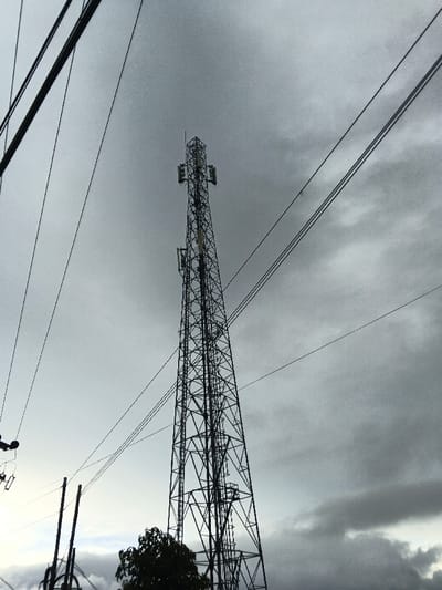 The image displays a low-angle, upward-looking view of a tall, dark-colored lattice telecommunications tower set against an overcast sky in Sindangjaya, Indonesia. The tower, centrally located, features multiple rectangular panel antennas mounted at various heights along its structure, with a lightning rod or antenna extending from its apex. Numerous parallel utility wires traverse the frame, crossing both the upper and middle sections of the image. On the far left, two dark utility poles with associated wiring and a small apparatus are partially visible. A portion of a dark-leaved tree occupies the bottom-center foreground, obscuring the base of the tower. The sky is predominantly gray with varying cloud densities, indicating overcast conditions. No individuals are present in the scene.