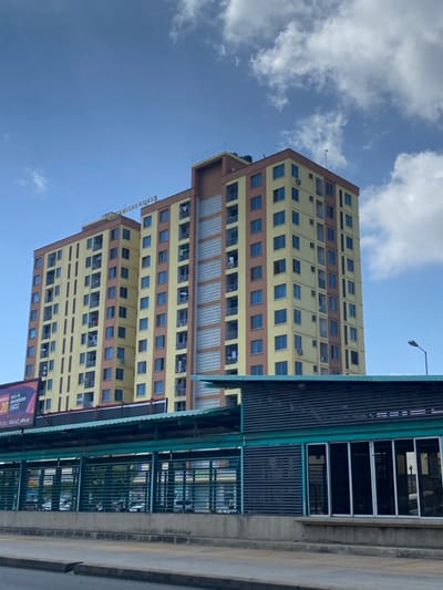 The image displays a tall, multi-story building dominating the left and central parts of the frame, set against a bright blue sky dotted with scattered white clouds. The building has a distinctive architectural style, featuring light yellow walls accented by horizontal brown bands that separate its many floors. Numerous rectangular windows, appearing dark or blue, are visible across the facade, with some balconies showing clothes hanging to dry, suggesting residential use. Partial text on the side of the building appears to read "Bird Residences."
In the foreground, on the right side, an elevated infrastructure component, possibly an overpass, walkway, or part of a public transport station, extends into the frame. This structure is built from concrete and features prominent green metal railings and dark slatted panels. A street light pole is visible above the building. At the bottom right, a small portion of a billboard is visible, displaying text that includes "20" and "EA CLC official."
The scene depicts a clear, sunny daytime. No specific human activity is actively taking place. The overall impression is that of an urban landscape, consistent with the provided context of Dar es Salaam, Tanzania, featuring modern architecture and infrastructure.