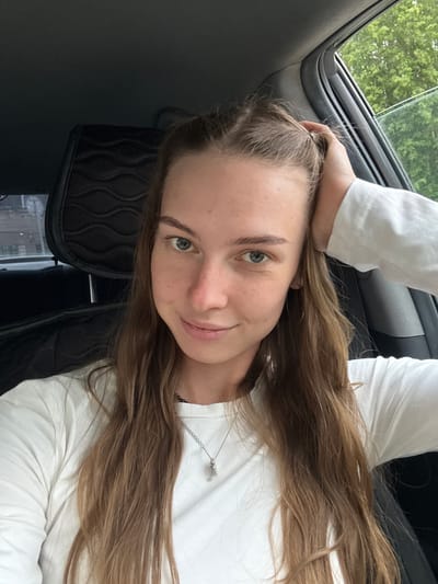 A young woman with long, light brown hair pulled back with a hand is seated in the driver's seat of a car. She is wearing a white long-sleeved shirt and a silver necklace with a key pendant. Her gaze is directed at the camera, and she has a slight smile on her face. The interior of the car is visible, including a black headrest with a patterned design. Outside the car window, green trees and foliage can be seen, indicating an outdoor setting, possibly on the outskirts of Perm, Russia, during daylight hours.