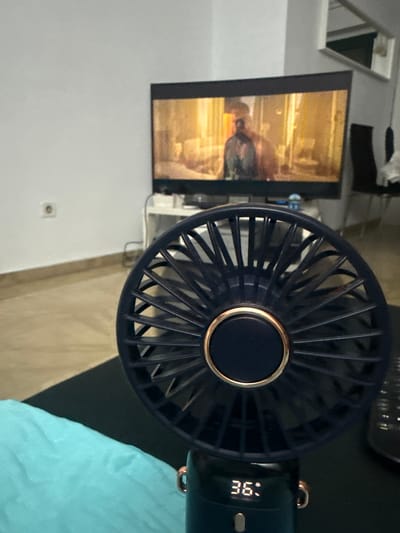 The image shows a close-up of a portable fan in a room. The fan is dark blue with a gold accent around the center. A digital display on the fan shows "36". To the left of the fan is a television screen displaying a still image of a person in a dimly lit, possibly historical, setting. The overall scene appears to be indoors, with a neutral-colored wall on the left and a textured surface, possibly a bed or couch covered with a light blue fabric, on the right. The lighting suggests it could be daytime or nighttime, but the interior setting is the primary focus. There are no overt indicators of weather or specific emotional cues. The fan and the television are the main objects, with the television displaying a static image rather than an active scene. The context of Valencia, Spain, doesn't provide specific visual cues within the image itself.