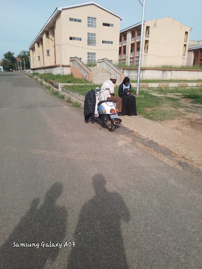 Two individuals are seated next to a parked scooter on an asphalt road in front of buildings. One person, a male wearing a light-colored striped shirt and a cap, is seated on the scooter. The other person, a female wearing a black hijab and a dark dress, is seated on the ground next to him. The scooter is light blue and white. In the background, there are two multi-story buildings with light-colored exteriors and windows. The environment appears to be an outdoor setting, possibly a campus or public area, with some greenery and paved walkways. The scene is captured under natural lighting, casting shadows on the road. The text "Samsung Galaxy A03" is present in the lower left corner of the image.