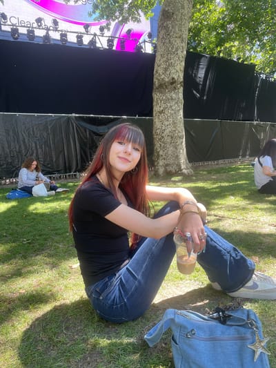 A young woman with long dark hair with red streaks and bangs is sitting cross-legged on a grassy area, holding a drink. She is wearing a black t-shirt and blue jeans, and several rings on her fingers. In the background, a stage with a black backdrop and large screens with pink lettering is visible. Other people are sitting on the grass in the distance. The scene appears to be outdoors in a park, likely at an event or festival. The event appears to be a concert or music festival, as suggested by the stage setup and the presence of a crowd.