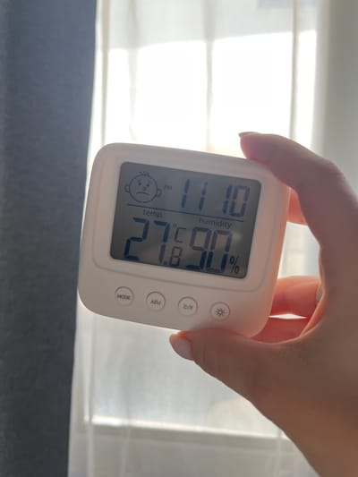 A white digital thermometer/hygrometer is held in a person's hand, with the device displaying a temperature of 27.8°C and a humidity of 90%. Above the temperature, a sad face icon is shown, possibly indicating the current conditions are not ideal. The time displayed is 11:10 PM. The background shows sheer curtains allowing bright light to pass through. The location is not discernible from the image.