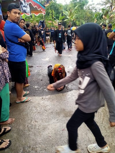 The scene depicts an outdoor event or performance occurring on a wet paved road in Mojogedang, Indonesia. In the immediate foreground, a young person wearing a black hijab, a grey long-sleeve shirt, black pants, and white Crocs walks from right to left. To the left, a man in a blue t-shirt, black cap, and dark shorts stands with crossed arms, looking forward. Directly behind him, another person is partially visible. Centrally located in the mid-ground, a person lies face down on the wet asphalt, wearing a distinctive, ornate traditional mask featuring a red face, prominent teeth, and long, shaggy brown and orange hair. Dark clothing is visible beneath the mask. In the background, a crowd of approximately 20-30 individuals, primarily men, are gathered and observing. Many are dressed in black t-shirts, some bearing white graphics. Tropical foliage, including banana trees, is visible behind the crowd. A temporary structure with a red, blue, and black banner featuring white text is erected on the left side of the background. One man in the mid-background walks barefoot, holding a red-tasseled object, and smiles towards the camera.