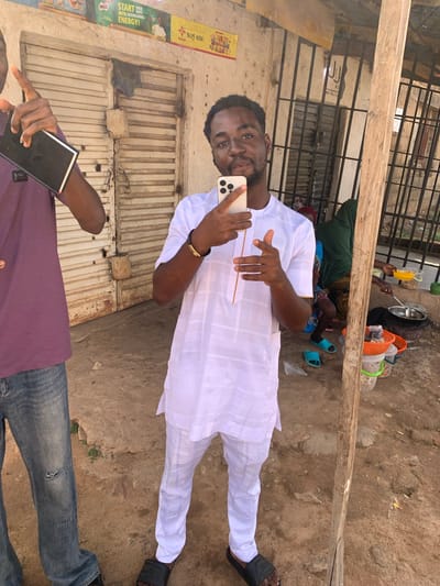 A medium shot shows a man in a white tunic and pants, holding a smartphone in front of him and gesturing with his other hand, in Jos, Nigeria. The man has dark skin and a short beard. He wears a gold bracelet.
Behind the man are metal roll-up doors and a vertical sign with the text "START THEIR DAYS WITH NOURISHING ENERGY!" A second man is visible at the bottom of the shot, wearing a purple shirt and jeans, also holding a device.
In the background, there are cooking utensils and people, one wearing a green headscarf, resting on the ground. The ground appears to be dirt or gravel. A wooden beam runs diagonally across the upper part of the image.