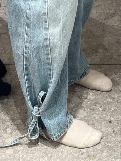 The image shows a close-up view of a person's legs wearing light blue denim jeans with a tie detail at the ankle. They are also wearing off-white socks. The floor appears to be made of speckled terrazzo tiles, and a shadow is cast on the floor to the right. The background is out of focus, but the overall environment suggests an indoor setting. There is no visible indication of the location being Berlin, Germany.
