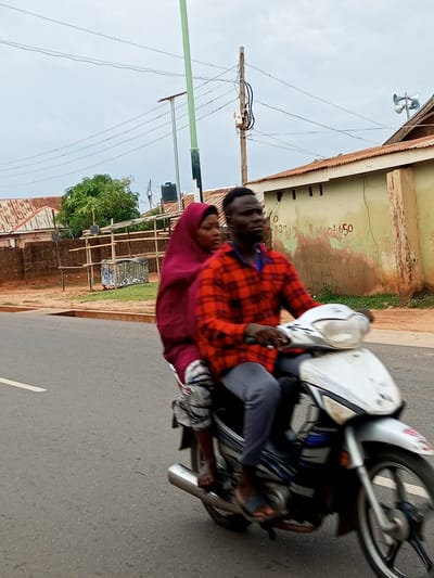 A man and a woman are riding a motorcycle on a paved road. The man is driving, wearing a red and black plaid shirt and grey pants. The woman is a passenger behind him, wearing a maroon headscarf and a patterned skirt. The motorcycle is white and silver with black accents.
The scene appears to be in Gombe, Nigeria. In the background, there are buildings with corrugated metal roofs and stucco walls. One building has a sign with red numbers that appear to read "350" or "650." There are also utility poles and overhead wires, typical of an urban or semi-urban environment. A large green tree is visible on the left side of the image. The sky is overcast, suggesting a cloudy or possibly rainy day. The overall lighting indicates it's likely daytime, but the overcast conditions mute the brightness. The subjects' expressions are not clearly discernible due to the motion and resolution. The activity is simply the two individuals traveling on the motorcycle.