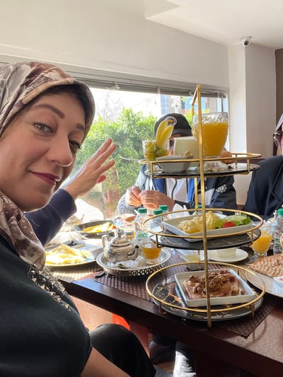 A woman with a patterned hijab and dark sweater is positioned in the foreground, taking a selfie, her right hand raised with palm facing the camera. She has green eyes and wears pink lipstick. Seated at a table with her are at least two other individuals; one man wearing a dark baseball cap with a logo and a blue and white jacket is visible in the mid-ground, appearing to interact with food.
The setting is an indoor restaurant or cafe in Meknes, Morocco, characterized by large windows in the background revealing green foliage and bright daylight. The ceiling is white, featuring a mounted white security camera in the upper right corner.
A prominent three-tier serving stand with gold-colored metal frames and black and gold marble-patterned plates occupies the center of the table. The top tier holds a glass of orange juice, a small white bowl of a light substance, and a glass with cut fruit (kiwi, grapes) and two banana slices. The middle tier displays a plate of scrambled eggs and a small side salad. The bottom tier features a plate of French toast or pancakes topped with nuts.
The dark brown wooden table is laden with various breakfast items, including a silver-colored Moroccan teapot on a tray with a glass of amber tea, plates of eggs, cheese, bread, and bottled water. The overall scene depicts a group partaking in a shared meal.