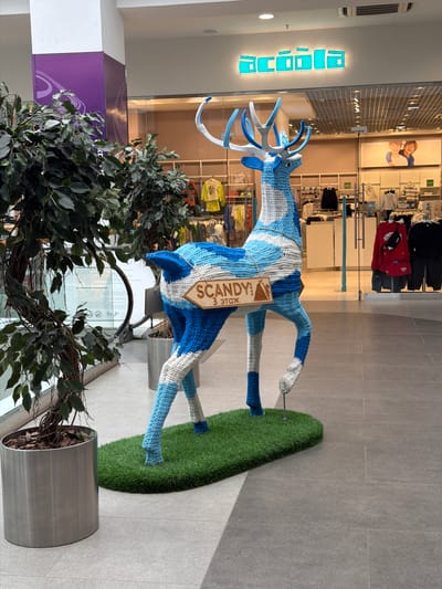 The image shows a large, crocheted sculpture of a deer, predominantly blue and white, standing on a patch of artificial grass. The deer sculpture is the main subject, positioned in the center of the frame. It has elaborate antlers, also crocheted in white and blue. A wooden tag hangs from its neck, displaying text in a diamond shape. Visible text on the tag reads "SCANDY PARK" and "3 ЭТАЖ" (3rd floor) in Cyrillic.
Behind the deer, there is a shop interior visible through a glass storefront. The shop appears to be a clothing store, with racks of clothes, mannequins, and shelving units. The lighting suggests an indoor environment, likely a shopping mall. A prominent turquoise illuminated sign with geometric shapes is mounted on a wall to the left of the deer, also displaying Cyrillic text which seems to be "Acoola".
In the foreground, to the left, is a large potted plant with dark green leaves and visible branches, partially obscuring the lower part of the deer sculpture and the shop. To the right of the deer, the floor is tiled with large grey tiles.
The image appears to be taken during the day, given the lighting conditions. There are no explicit indicators of weather or time of day beyond the indoor lighting. The scene suggests a public space, likely a mall in Izhevsk, Russia, where the deer sculpture is an art installation or decoration. The overall impression is one of a festive or welcoming display within a commercial setting.