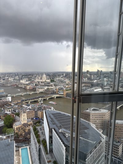 This high-angle, wide shot captures a panoramic view of a vast urban landscape, identifiable as London, United Kingdom, under an overcast sky. The photograph appears to be taken from a significant height, likely an observation deck of a skyscraper, looking down through large window panels that show reflections of the gray, cloudy sky.
The prominent feature of the scene is the River Thames, which snakes horizontally across the mid-ground, traversed by multiple bridges. Several boats, including what appears to be a larger passenger vessel, can be seen navigating the murky brown waters of the river. The cityscape sprawls densely on both banks, showcasing a mix of architectural styles. Modern glass-clad high-rises stand alongside older, more traditional brick and stone buildings, some with distinctive rooftops and intricate detailing. In the far distance on the left, the dome of a significant landmark, possibly St Paul's Cathedral, is faintly visible amidst the urban sprawl.
In the immediate foreground and below the vantage point, parts of the building from which the photo was taken are visible, including steel window frames and structural elements. Adjacent buildings reveal rooftop details, including a striking turquoise swimming pool on a lower rooftop terrace. Further down, outdoor seating areas and pedestrian zones are discernible, suggesting active public spaces, though no individual people are clearly identifiable. The weather is distinctly cloudy and gray, indicating an overcast day without direct sunlight, contributing to a muted and slightly somber atmosphere across the metropolis. No discernible text is visible on any buildings or objects within the frame.