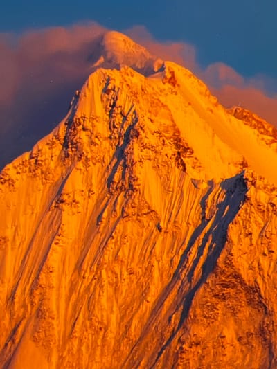 The image is a close-up shot of a snow-covered mountain peak illuminated by warm, orange light, likely from a sunrise or sunset. The mountain is rugged with visible texture and shadows, suggesting steep slopes and rocky outcrops beneath the snow. Wisps of clouds, tinged with pink and orange, are visible against a deep blue sky in the upper portion of the image. The overall impression is one of a dramatic and majestic natural landscape. There is no indication of Zichem, Belgium, or any urban environment in this image.