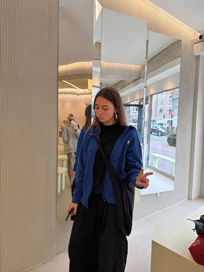 In Amsterdam, Netherlands, a young woman with long brown hair is captured in a selfie, her face angled downwards and slightly to the side, her lips pursed. She wears a black t-shirt beneath a vibrant royal blue zippered hoodie, and loose black pants. A black crossbody bag is slung over her right shoulder. Her left hand is raised, fingers extended in a peace sign, while her right hand holds a smartphone. The background features a textured, ribbed wall to the left and a floor-to-ceiling mirror that reflects the scene, including another person in the background wearing a light grey hoodie and patterned pants, also taking a selfie. The mirror's surface is slightly warped in places, reflecting the ambient lighting and the exterior view of a street with buildings and a parked car. To the right, a white counter displays a red bag with white lettering. The overall environment appears to be a retail space or fitting room with clean, modern aesthetics.