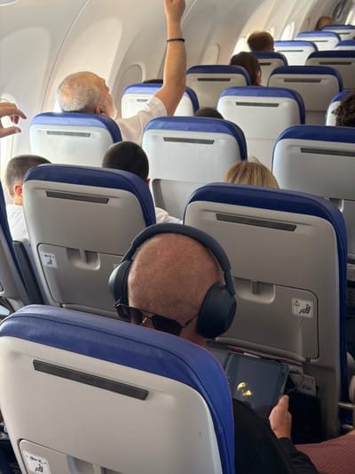 The image shows an interior view of an airplane cabin, with rows of seats visible. The perspective is from a person sitting in an aisle seat, looking forward. Several passengers are present, mostly seen from the back or side. One passenger in the foreground is wearing headphones and sunglasses, and is looking at a tablet device. Another passenger is visible in the aisle in the mid-ground, their arm outstretched. The seats are upholstered in blue and white. There are safety instruction symbols visible on the back of some seats. The lighting suggests it is daytime, and the overall mood appears relaxed, with passengers engaged in their own activities. Visible text includes "RECARO" on the back of a seat, indicating the seat manufacturer. The image is captured in a landscape orientation.