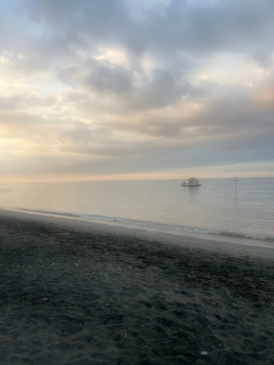 The image shows a serene beach scene with dark sand in the foreground and the calm ocean stretching towards the horizon. The sky is filled with overcast clouds, tinged with soft yellow and grey hues, suggesting either dawn or dusk. In the distance, a small, isolated hut floats on the water, appearing to be a simple dwelling or a small boat. A few figures are visible on or around the hut. The overall atmosphere is tranquil and slightly hazy. The location is described as Gerokgak, Indonesia.