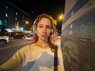 A woman with curly hair and headphones is pictured in a selfie. The background suggests she is in Novi Sad, Serbia, due to the presence of cars and buildings characteristic of the area. A street with traffic can be seen. Buildings with signage are visible.