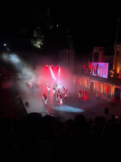 The image depicts a night performance taking place in an ancient Roman amphitheater in Plovdiv, Bulgaria. A group of performers, dressed in traditional white and red costumes with headdresses, are on stage, illuminated by red and white stage lights and smoke effects. Some performers are dancing, while others appear to be posing. A large screen behind the performers displays images of people in similar attire. The amphitheater's seating area, filled with an audience, is visible in the background and foreground, mostly silhouetted against the stage lights. The architectural elements of the ancient structure, including stone columns and tiers of seating, are illuminated by warm orange and red lights.