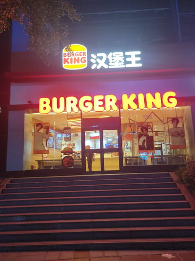 This vertically oriented image captures the illuminated facade of a Burger King restaurant at night, likely in Shapingba District, China. The scene is dominated by bright neon signage against a dark backdrop, indicating the time of day.

On the left, a large, vibrant yellow and red neon sign spells "BURGER KING" vertically. Adjacent to it, another neon sign in white displays the Chinese characters "汉堡王" (Hànbǎo Wáng), which translates to Burger King. Below these, a smaller, oval-shaped sign features the classic Burger King logo. A leafy tree partially obstructs the view on the far left.

The center of the image shows the glass-fronted entrance of the restaurant, revealing a brightly lit interior. Several large promotional posters are visible through the windows, primarily featuring individuals (likely models) holding or interacting with food items. Other menu displays and a general view of the restaurant's inside are also discernible. A faint, blurred figure of a person can be seen through the glass inside the restaurant.

To the right of the entrance, a wide, dark staircase with prominent vertical risers ascends out of frame. A wall with dark vertical panels runs alongside these stairs. The overall impression is an active, open fast-food establishment at night, drawing attention with its prominent and colorful lighting.