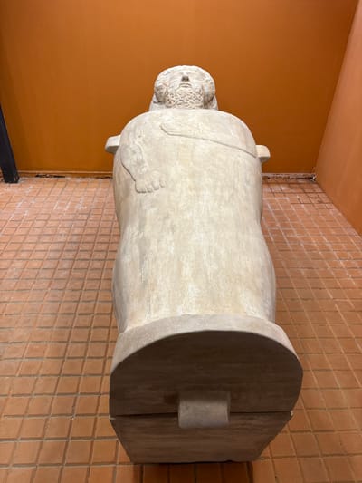 A large, light-colored anthropomorphic sarcophagus is displayed indoors, oriented with its head facing away from the viewer and slightly upwards. The sarcophagus depicts a male figure with a bearded face gazing upward, stylized hair or headdress, and arms bent with hands resting on the chest. It rests on a flat, rectangular base. The immediate environment features solid, unadorned orange-ochre colored walls that meet in a corner to the right of the sarcophagus. The floor is covered with small, square, reddish-brown tiles laid in a grid pattern. A dark vertical element is partially visible on the far left. This object is characteristic of the Phoenician/Punic anthropomorphic sarcophagi found in the region and is specifically located within the Museo de Cádiz in Cádiz, Spain.